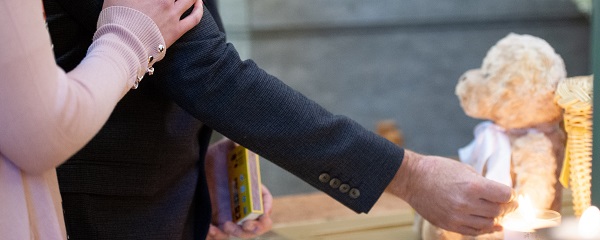 Man lighting a candle with teddy bear in background and woman's hand on his arm