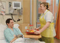 A patient is sitting up in a hospital bed as one of the hospital staff gives them their food on a tray